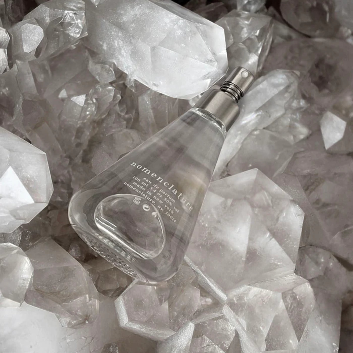 Nomenclature - adr_ett Sprayflakon mit transparentem Glas und silbernem Zerstäuber auf Kristallsteinen liegend