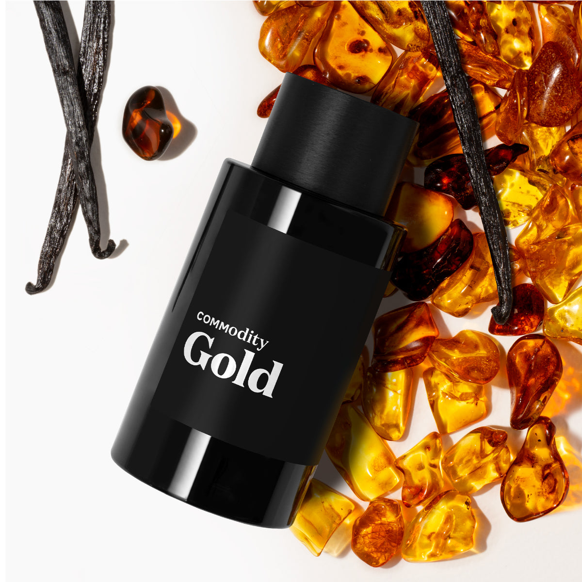 Commodity - Gold Parfumflakon in mattschwarzem Design mit Bernsteinsteinen und Vanilleschoten auf hellem Hintergrund