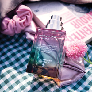 The Different Company - Love is coming...Chapter II Parfumflakon in mehrfarbigem Glas auf kariertem Tuch mit Buch und Blume
