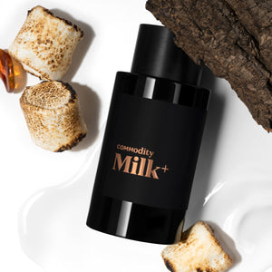 Commodity - Milk+ schwarzer Flakon mit kupferfarbenem Schriftzug neben gerösteten Marshmallows und Holzstück auf weißem Grund