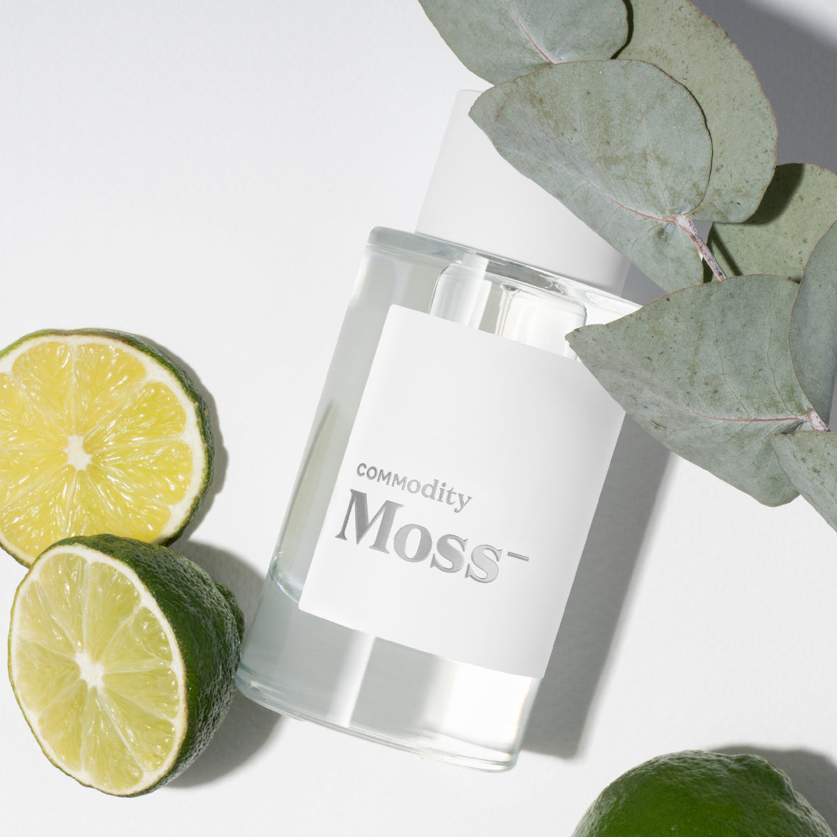 Commodity - Moss- Duftflakon mit klarem weißen Etikett, Limettenscheiben und grünen Blättern auf hellem Untergrund