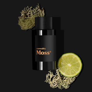 Commodity - Moss+ schwarzer Flakon mit kupferfarbenem Schriftzug umgeben von Limette und Moos vor schwarzem Hintergrund