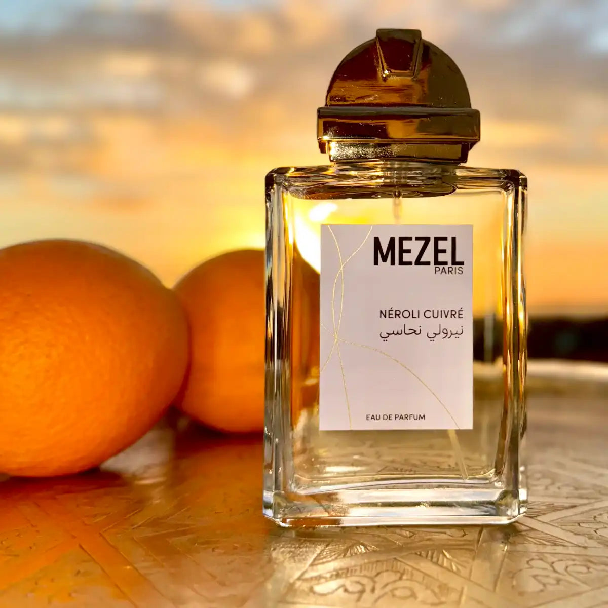 Mezel Paris - Néroli Cuivré Parfumflakon mit goldenem Verschluss vor Sonnenuntergang und zwei Orangen im Hintergrund