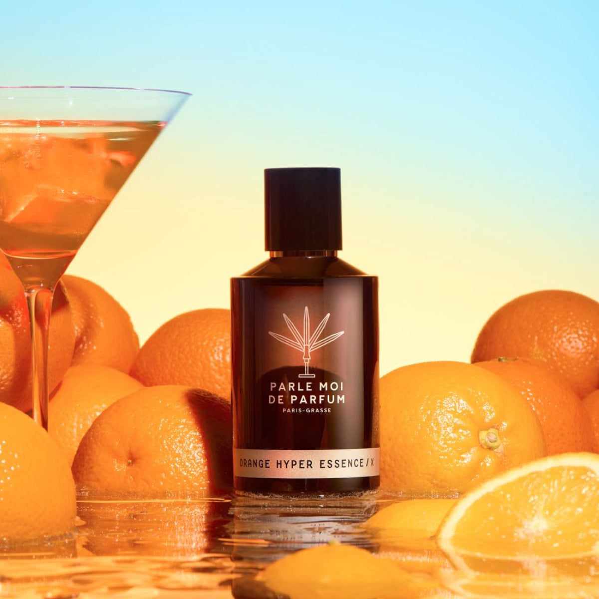 Parle Moi de Parfum - Orange Hyper Essence / X Flakon umgeben von sonnigen Orangen und Cocktailglas vor hellem Himmel