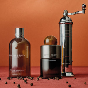 Molton Brown - Re-Charge Black Pepper Set mit Duschgel, Duftflakon mit rundem Verschluss und silberner Pfeffermühle vor orangefarbenem Hintergrund