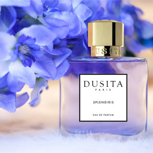 Dusita - Splendiris Parfumflakon mit zartviolettem Inhalt und goldfarbenem Verschluss vor blauen Blüten auf hellem Hintergrund