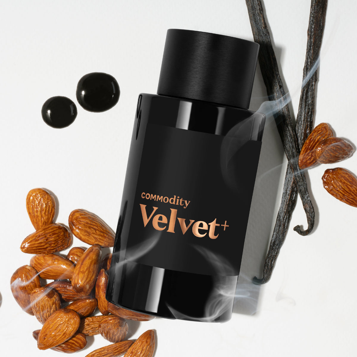 Commodity - Velvet+ schwarzer Flakon mit kupferfarbenem Schriftzug umgeben von Vanilleschoten und Mandeln auf weißem Hintergrund