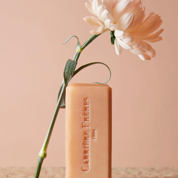 Carrière Frères - Florae - Seife als zartrosafarbener Block mit geprägtem Schriftzug und zarter Blüte vor pastellfarbenem Hintergrund