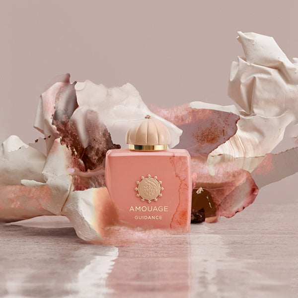 Amouage - Odyssey Collection - Guidance Flakon in zartem Rosa mit goldfarbenem Emblem vor künstlerischem Hintergrund