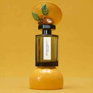L'Artisan Parfumeur - La Collection - Histoire d'Orangers Parfumflakon mit kunstvollem Orangen-Deckel vor gelbem Hintergrund