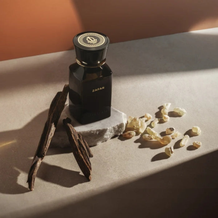 Omanluxury - Zafar Parfüm in dunklem Glas mit goldener Schrift, umgeben von Oudholz und Weihrauch vor schlichtem Hintergrund